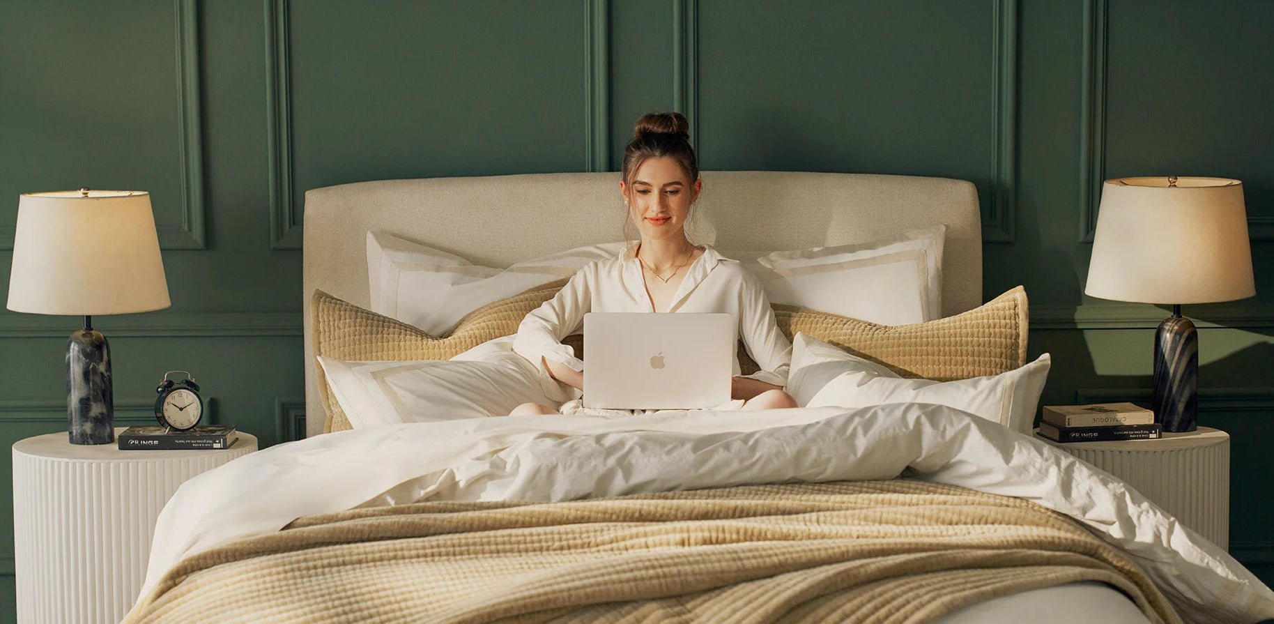 Woman sitting on a bed with a laptop, surrounded by soft lighting in a bedroom setting.