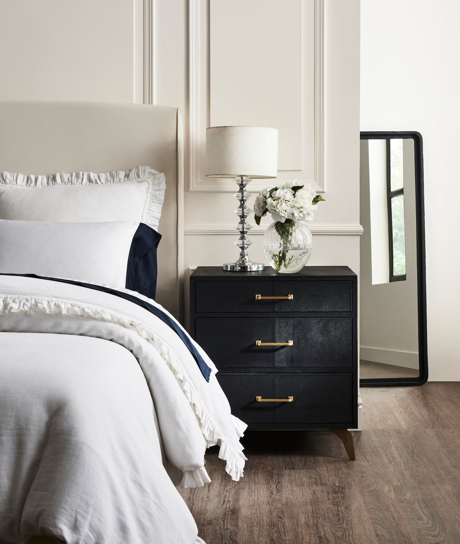 FN-5200-262028_1|Sleek black three-drawer nightstand with gold bar pulls & modern tapered legs beside a cozy white bed setup.