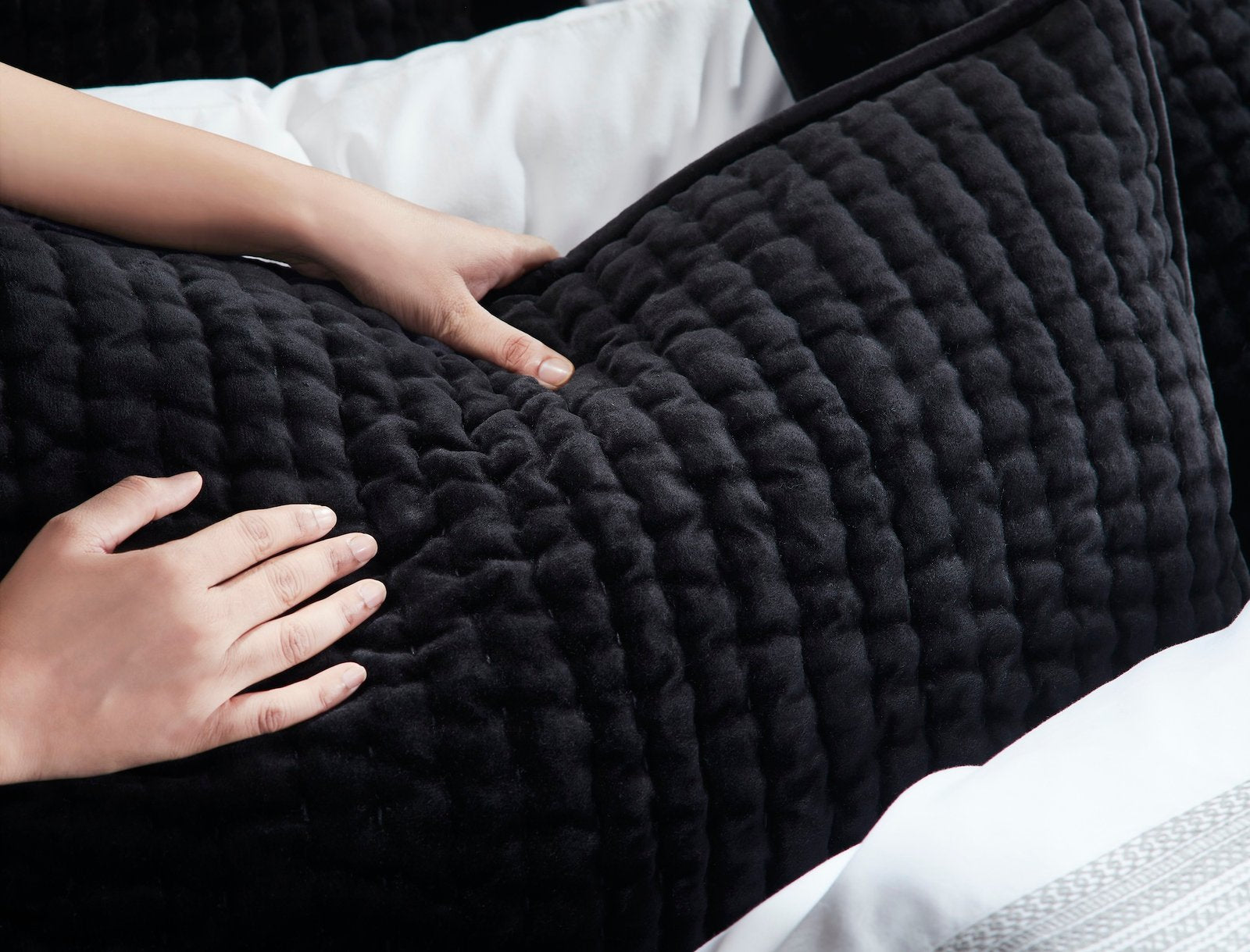 QT-361-9496_6|Hand gently touching the rich, textured fabric of a soft black quilted pillow, emphasizing luxurious comfort and quality.